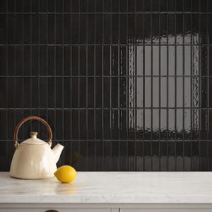 Op de afbeelding: Een crèmekleurige keramische theepot met een bruin gevlochten handvat staat naast een enkele heldergele citroen. De achtergrond bestaat uit een muur van glanzende, verticale zwarte rechthoekige tegels in een gestapeld patroon.