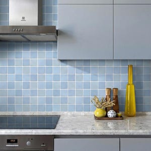 Könnte beinhalten: Eine moderne Küche mit hellblauen quadratischen Fliesen, einer Edelstahl-Dunstabzugshaube und hellgrauen Schränken. Eine Marmorarbeitsplatte zeigt dekorative Gegenstände, darunter eine gelbe Vase und hölzerne Gewürzbehälter.