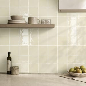 May include: A kitchen backsplash featuring square, glossy, cream-colored tiles with visible grout lines. A wooden shelf holds a stack of plates, a ceramic mug, and two small glasses. A bottle of oil and a bowl of pears sit on the countertop.