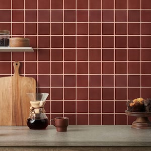 May include: A kitchen scene with a red-brown tiled backsplash. A wooden cutting board, a glass coffee maker, and a brown mug sit on a gray countertop. A small wooden shelf holds a jar and a wooden bowl. A cupcake sits on a wooden cake stand.