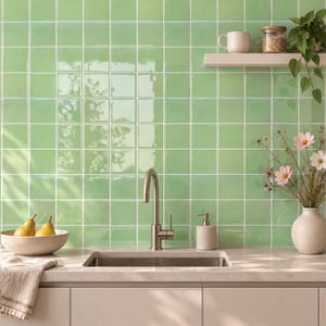 May include: A kitchen backsplash featuring glossy, square light green tiles with white grout lines. The setup includes a stainless steel faucet, a beige soap dispenser, a bowl with two yellow pears, and a vase with pink flowers on a countertop.