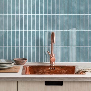 May include: A kitchen setup featuring a rectangular hammered copper sink with a matching copper faucet. The backsplash consists of vertical, glossy, teal-coloured rectangular tiles with white grout lines. A wooden cutting board sits nearby.