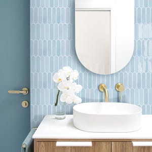 May include: A white porcelain bathroom sink with a gold tap and a white orchid in a glass vase. The sink is set against a wall tiled with light blue, arched tiles.