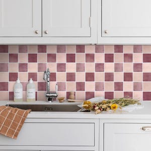 Puede incluir: Una cocina con un fregadero de acero inoxidable y armarios blancos. El salpicadero presenta un patrón a cuadros de cuadrados rosas y malvas. Dos botellas blancas y un paño de cocina están en la encimera.