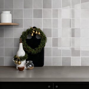 Könnte beinhalten: Eine Küchenszene mit einer grauen Arbeitsplatte und einem schwarzen Unterschrank. Eine weiße Flasche, ein kleines Glas und eine Holzschale stehen auf der Theke. Ein schwarzes Schneidebrett ist mit einem grünen Kranz und goldenen Glocken verziert. Der Hintergrund ist eine geflieste Wand.