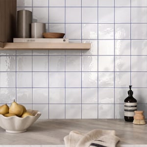 May include: A wall covered in square white ceramic tiles with dark grout lines. A wooden shelf holds two cylindrical containers and a small wooden bowl. A white bowl filled with pears, a dark glass bottle, and a wooden brush sit on the counter.