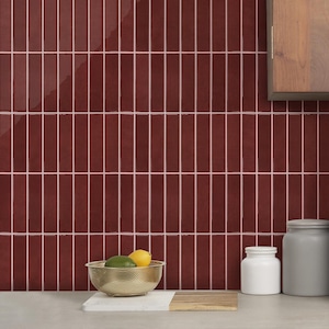 Può includere: Una parete rivestita con piastrelle verticali rettangolari di colore rosso scuro lucido con sottili fughe bianche. Una ciotola in rete metallica dorata contenente un lime verde e un limone giallo poggia su un tagliere bianco e legno.