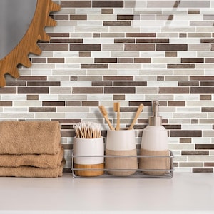 Könnte beinhalten: Badezimmerdekoration mit einem Stapel gefalteter brauner Handtücher, einem Keramikbehälter mit Wattestäbchen, Zahnbürsten und einem Seifenspender. Der Hintergrund ist eine Mosaikfliesenwand mit braunen, weißen und grauen rechteckigen Fliesen.