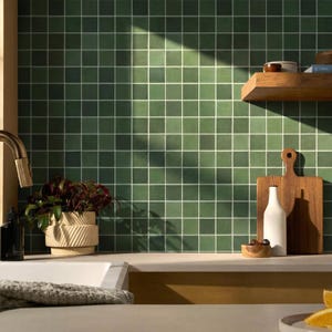 Könnte beinhalten: Kücheninterieur mit grüner Fliesenwand und weißer Fuge. Ein Holzregal hält kleine Schalen. Eine weiße Flasche und ein Holzschneidebrett stehen auf der Theke. Eine Topfpflanze und ein Waschbecken befinden sich im Vordergrund. Sonnenlicht fällt in den Raum.