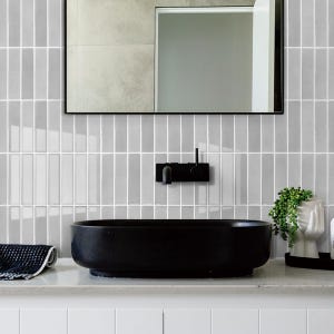Peut inclure: Une salle de bain moderne avec un lavabo ovale noir, un robinet noir et un grand miroir. Les murs sont recouverts de carreaux verticaux gris clair. Une petite plante dans un vase blanc est sur le comptoir.