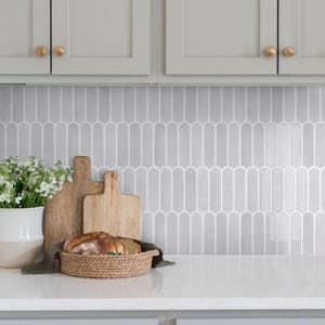 Peut inclure: Scène de cuisine avec un plan de travail blanc, une crédence grise et des meubles gris clair avec des boutons dorés. Un panier en osier contient du pain, des planches à découper en bois et un vase de fleurs blanches.