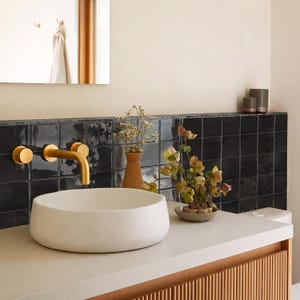May include: A white circular vessel sink sits on a light-colored countertop next to a gold wall-mounted faucet. Behind the sink is a black square-tiled backsplash, a ribbed brown vase with dried flowers, and a small bowl with green floral stems.