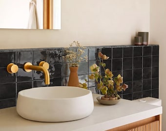May include: A white circular vessel sink sits on a light-colored countertop next to a gold wall-mounted faucet. Behind the sink is a black square-tiled backsplash, a ribbed brown vase with dried flowers, and a small bowl with green floral stems.