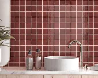 May include: A bathroom vanity area featuring a white cylindrical vessel sink and a tall, curved chrome faucet. The wall behind the sink is covered in square, glossy, deep red tiles with light-colored grout lines, set above a light wood cabinet.