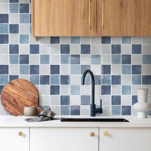 Puede incluir: Una cocina con un salpicadero de baldosas cuadradas en tonos azules y grises. Un mueble de madera con tiradores dorados está encima de las baldosas. Un grifo negro, una tabla de cortar de madera y una encimera blanca también son visibles.