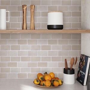 Może przedstawiać: Okładzina ścienna w kuchni z beżowymi płytkami typu metro. Drewniana półka z białym ceramicznym słojem z czarną pokrywką, dwoma drewnianymi młynkami do pieprzu i białym kubkiem. Miska z pomarańczowymi kaki znajduje się na blacie pod półką. Biały ceramiczny pojemnik na sztućce z czarną górą zawiera drewniane narzędzia kuchenne. Książka kucharska zatytułowana "Sous Vide at Home" leży na blacie.