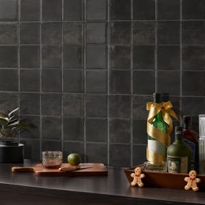 May include: Dark, glossy square tiles form a backsplash. A wooden board holds a lime, glass, and knife. Bottles of spirits are adorned with gold ribbon. Gingerbread men sit on a wooden tray.