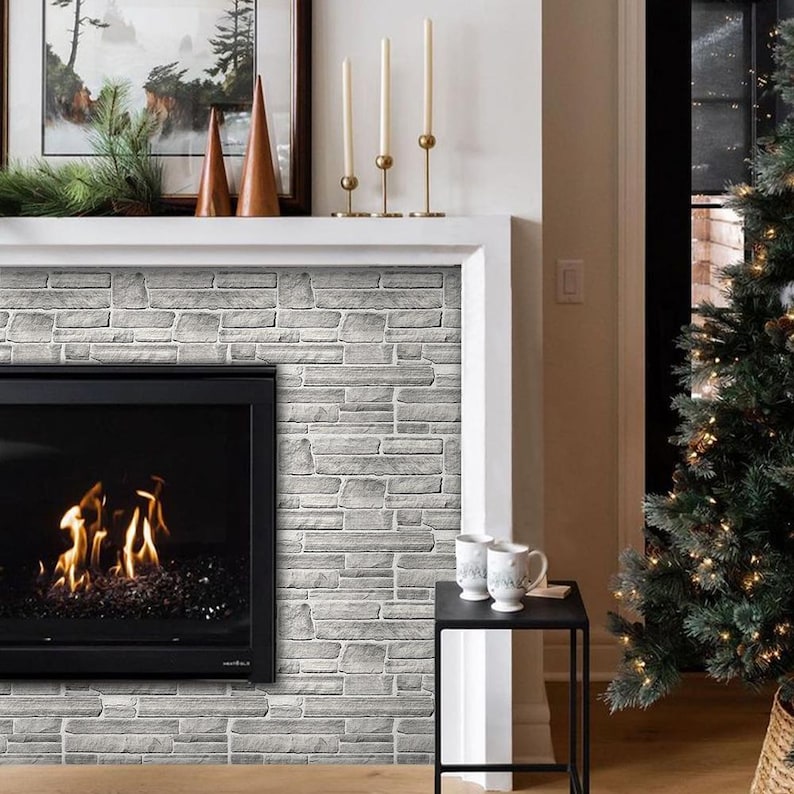 May include: A cozy living room scene featuring a fireplace with a gray brick-style accent wall. The fireplace has a black frame and a fire burning inside. A Christmas tree with lights stands to the right. Two mugs sit on a small black table.