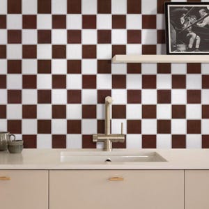 May include: A kitchen with a brown and white chequered tile backsplash. A stainless steel tap is above a white sink. A framed black and white photo hangs on the wall. Beige cabinets and a countertop complete the scene.