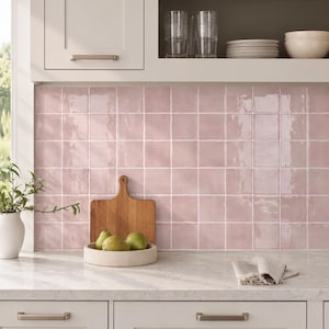 May include: A kitchen backsplash featuring square, glossy, light pink ceramic tiles with a subtle wavy texture. The tiles are arranged in a grid pattern with white grout lines, set against white cabinetry and a white marble countertop.