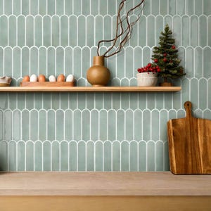 May include: A kitchen scene with a light green tiled wall and a wooden shelf. The shelf holds a small Christmas tree, a bowl of red berries, a brown vase, and a wooden egg holder with eggs. A wooden cutting board rests on the counter.