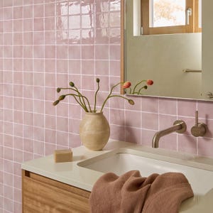 May include: A bathroom vanity area featuring glossy light pink square wall tiles, a white countertop with a built-in sink, a beige ceramic vase with flower buds, a rectangular bar of soap, and a textured dusty rose towel draped over the edge.