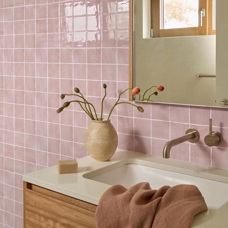 May include: A bathroom vanity area featuring glossy light pink square wall tiles, a white countertop with a built-in sink, a beige ceramic vase with flower buds, a rectangular bar of soap, and a textured dusty rose towel draped over the edge.