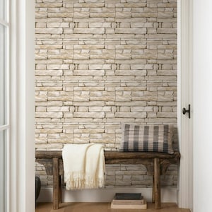 May include: A rustic wooden bench sits against a wall covered in light beige stone-look wallpaper. A cream-coloured fringed throw blanket is draped over the bench, accompanied by a grey and beige plaid rectangular pillow and two stacked books.