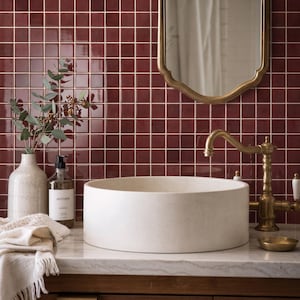 May include: A round, off-white stone vessel sink sits on a marble countertop in front of a deep red square-tiled wall. A brass faucet, a clear soap dispenser, a textured ceramic vase with eucalyptus branches, and a white fringed cloth are present.