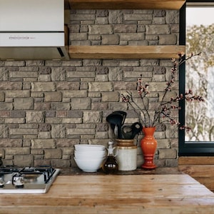 May include: A kitchen countertop with a brown wood surface. A gas stove with a black top and silver trim is on the left side of the counter. Three white bowls are stacked on the right side of the counter. A brown ceramic jar with a lid is next to the bowls. A clear glass bottle with a cork stopper is next to the jar. A red vase with a branch of dried flowers is on the counter. The background is a wall with a brown brick pattern.