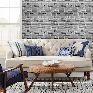 May include: A white tufted sofa with blue and white patterned throw pillows sits in front of a wall covered in a faux brick wallpaper. A woven wicker coffee table sits in front of the sofa.