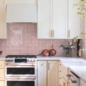 May include: A kitchen interior featuring white cabinets with gold handles, a white double oven range with copper accents, and a pink square tile backsplash. A white range hood is mounted above the stove, and a copper pan sits on the counter.