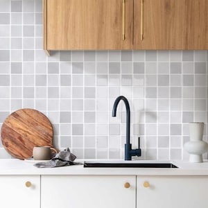 May include: A modern kitchen with a white countertop, black tap, and sink. The backsplash is tiled with small, square, light grey tiles. A wooden chopping board, a mug, and a vase are on the counter. Wooden cabinets are above.