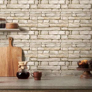 May include: A kitchen setting with a light stone wall. A wooden chopping board, a glass coffee maker, and a brown mug are on a grey countertop. A shelf holds a jar and a wooden bowl. A wooden cake stand holds muffins.