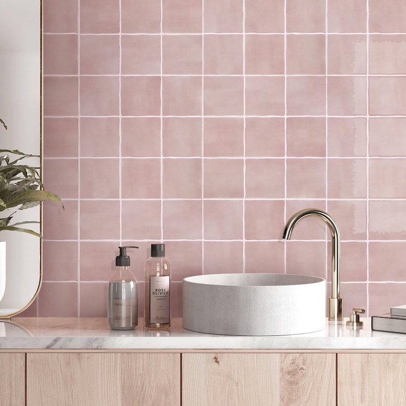 May include: A wall covered in square, dusty pink ceramic tiles with white grout lines. A white, cylindrical vessel sink sits on a light wood vanity next to a tall, curved gold faucet, a clear soap dispenser, and a clear bottle with a label.