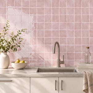 May include: A kitchen backsplash featuring square, glossy, light pink ceramic tiles with white grout lines. The wall is paired with a stainless steel gooseneck faucet, a white ceramic vase with white flowers, a bowl of pears, and a glass bottle.