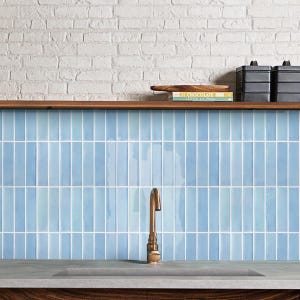 Op de afbeelding: Keukeninterieur met een lichtblauwe betegelde achterwand en een messing kraan. De tegels zijn verticaal gerangschikt. Een houten plank bevat boeken en zwarte bakken. De achtergrond is een witte bakstenen muur.