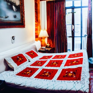 Könnte beinhalten: Ein Bett mit weißem Kopfteil und einer gemusterten Steppdecke. Die Steppdecke zeigt rote Quadrate mit floralen Designs auf weißem Hintergrund mit einem Wirbelmuster. Zwei passende rote Kissenbezüge sind sichtbar.