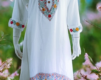 May include: White dress with colorful floral embroidery on the neckline, cuffs, and hem. The dress has a V-neckline with wooden buttons and three-quarter sleeves. The embroidery features red, yellow, and green flowers and leaves. The dress is displayed on a mannequin.