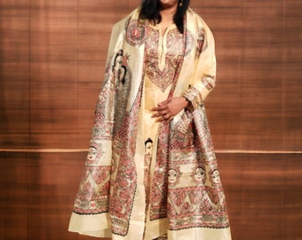 May include: A cream-colored ensemble featuring a tunic, pants, and a shawl, adorned with intricate red, pink, and black artwork. The outfit includes a long-sleeved tunic with detailed embroidery, matching pants, and a shawl with a repeating pattern of faces and floral designs.