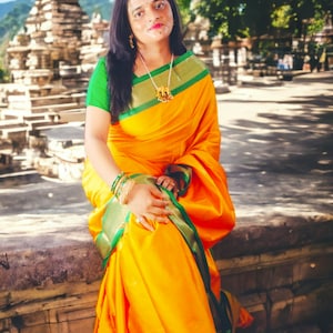 Op de afbeelding: Een vrouw in een felgele sari met groene en gouden randen, en een groene blouse. Ze draagt sieraden, waaronder een ketting en armbanden. De achtergrond toont een tempel en groene bladeren.