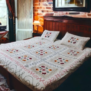Könnte beinhalten: Ein Bett mit einem dunklen Holz-Kopfteil und einer cremefarbenen, bestickten Steppdecke. Die Steppdecke weist ein sich wiederholendes geometrisches Muster mit roten, grünen und orangen Akzenten auf. Zwei passende Kissen liegen auf dem Bett. Eine Lampe steht auf einem Nachttisch.