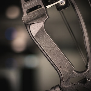 May include: Close-up of a black compound bow with a textured grip and the Hoyt logo. The bow has a black and silver design with a deer head logo.