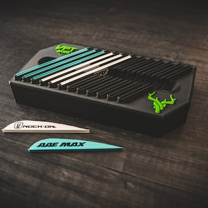 May include: A black arrow fletching jig with a green deer head logo. The jig has a series of slots for aligning fletching. Two fletching, one white with "nock on" text and one blue with "axe max" text, are shown on a wooden surface.