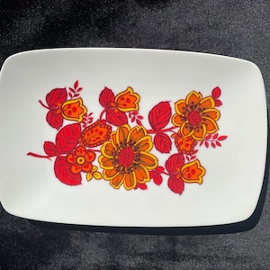 May include: A white rectangular serving dish with a floral design. The flowers are red, orange, and yellow with green leaves.