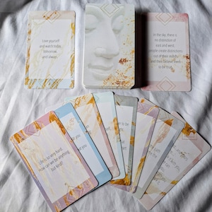 May include: Oracle cards with motivational quotes and geometric patterns in pink, gold, and blue. A white Buddha head statue is also visible. The cards are designed for spiritual guidance and self-reflection. The cards are approximately 12 cm in length.