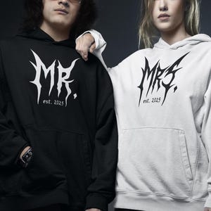 May include: Two people wearing black and white hoodies with the text "MR." and "MRS." in a stylized font, with "est. 2025" below. The person on the left wears black pants, and the person on the right wears a black skirt and fishnet stockings.