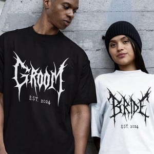Puede incluir: Dos camisetas a juego con texto gótico. Una camiseta es negra con "GROOM EST. 2024", la otra blanca con "BRIDE EST. 2024". Las camisetas presentan una fuente puntiaguda. Están de pie contra una pared de hormigón.