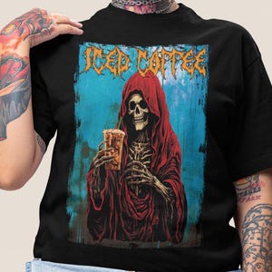 May include: Black t-shirt featuring a graphic of a skeleton in a red hooded robe holding an iced coffee. The words "Iced Coffee" are printed above the skeleton in orange, stylized lettering. The background is blue.