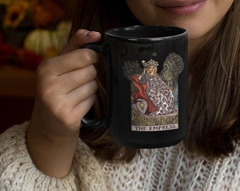 May include: Black ceramic mug featuring the Empress tarot card design. The illustration depicts a queen in a red and white gown, with the words "THE EMPRESS" below. The mug is held by a person wearing a cream-colored sweater.